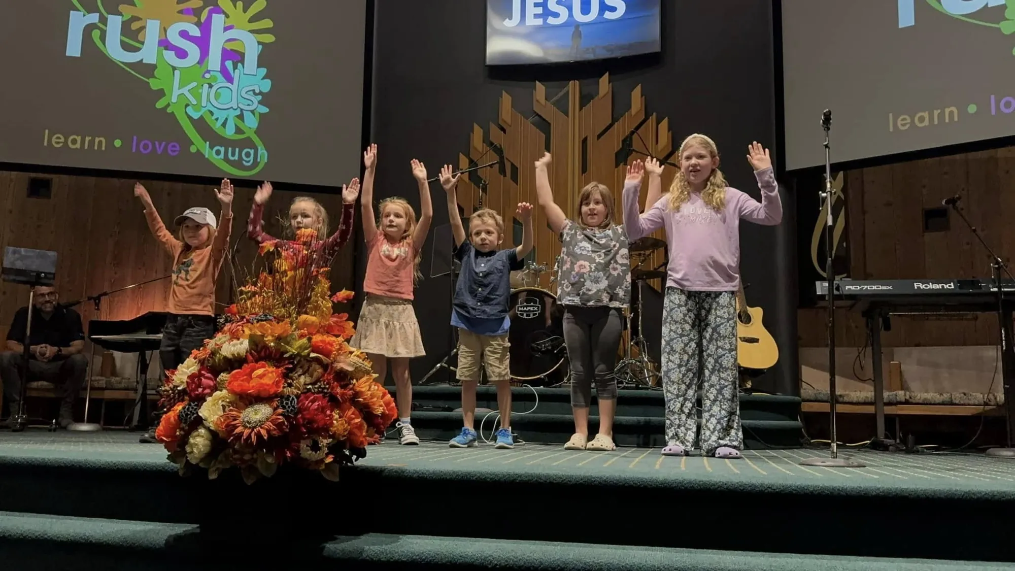 Children raising their hands on stage during a Rush Kids service, with "rush kids — learn love laugh" on the screen behind them